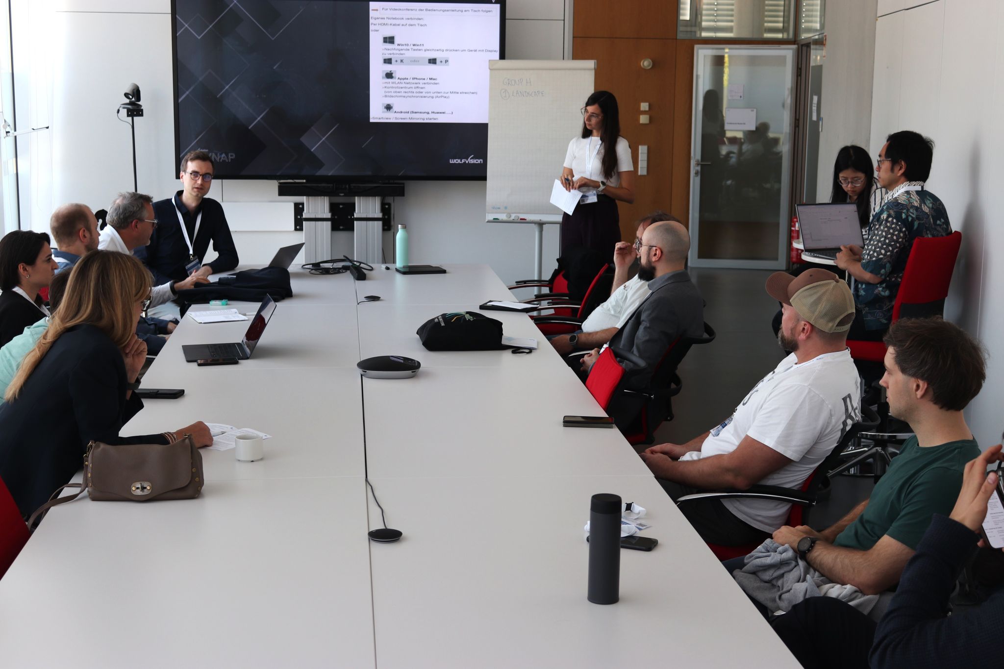 Participants gathered around a table during the Group H discussion, with a moderator standing beside a flipchart