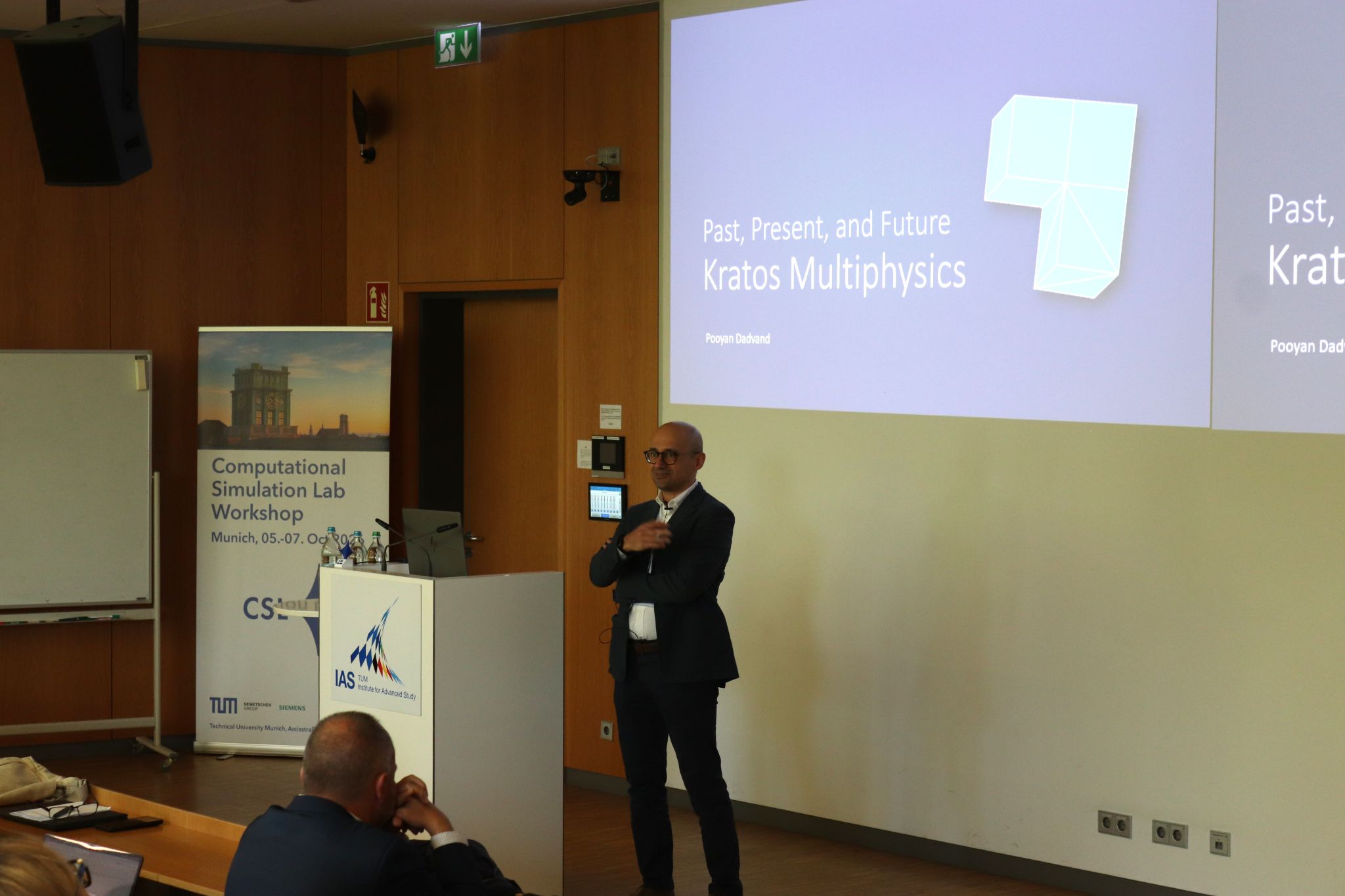 Pooyan Dadvand speaking beside a projected title slide about the past, present and future of Kratos Multiphysics