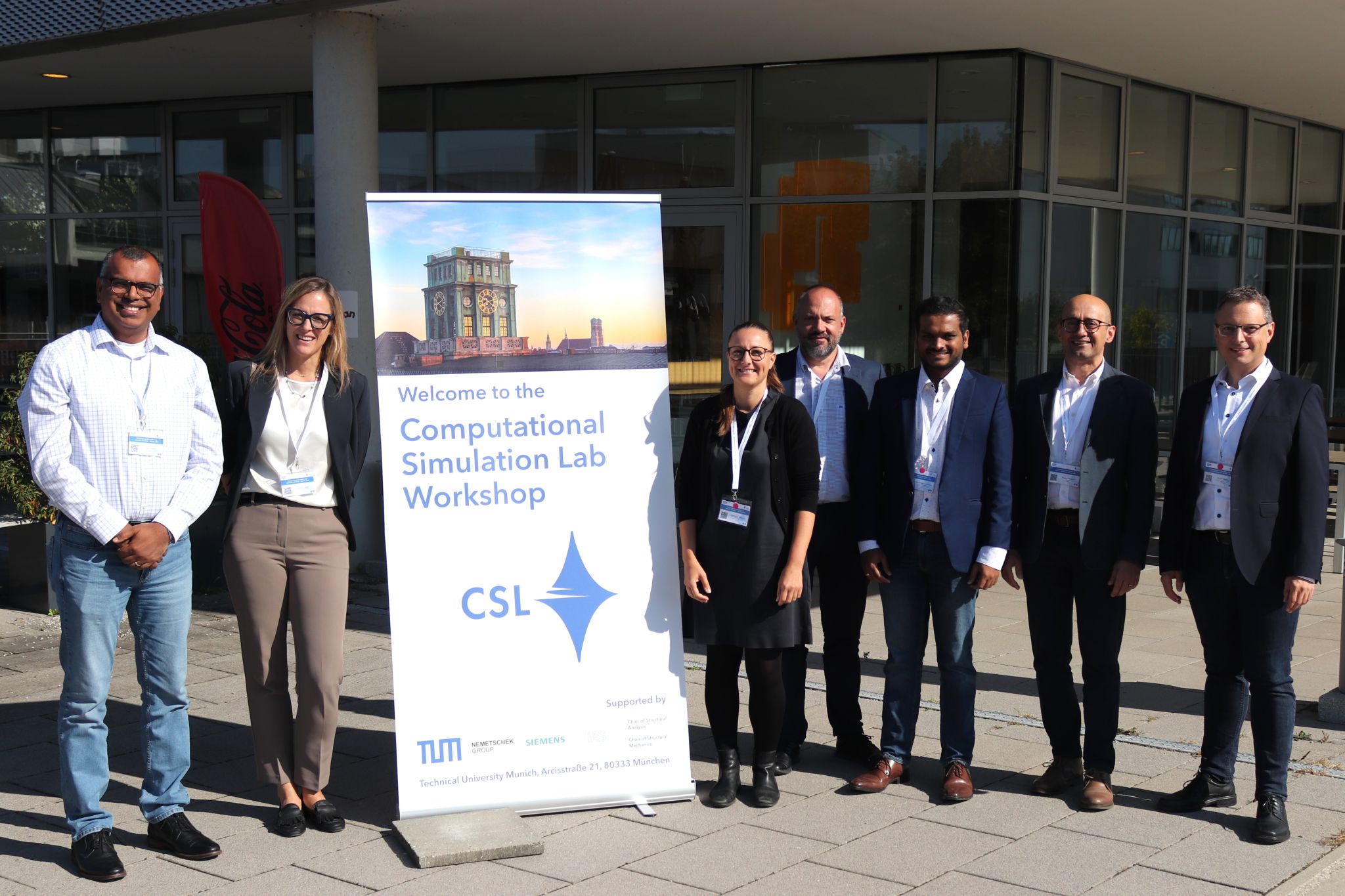 Seven members of the CSL workshop community standing beside the event welcome banner outside the venue
